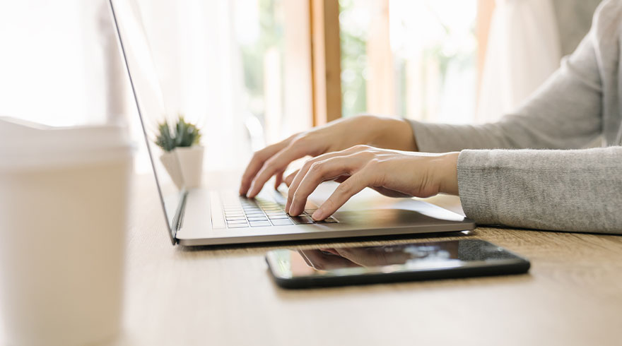 Een persoon typt op een laptop naast een telefoon op een houten tafel. Een kleine plant is zichtbaar op de achtergrond.
