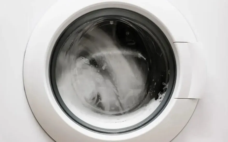 Een close-up van een draaiende wasmachine. Door de glazen deur is het water en schuim zichtbaar, wat de wasbeurt weergeeft. De witte deur en het omringende paneel contrasteren met de donkere binnenkant.