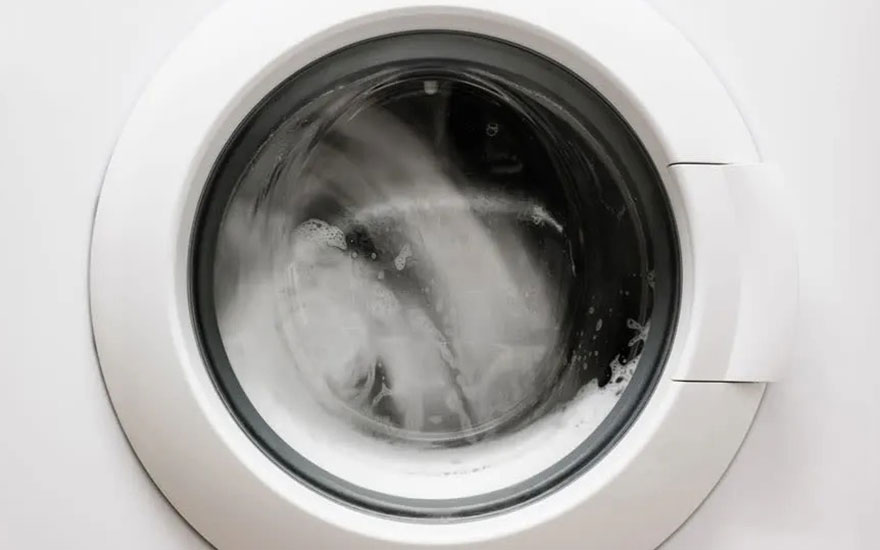 A close-up of a washing machine in motion. Through the glass door, one can see the water and suds, indicating it's mid-cycle. The white door and surrounding panel contrast with the dark interior.