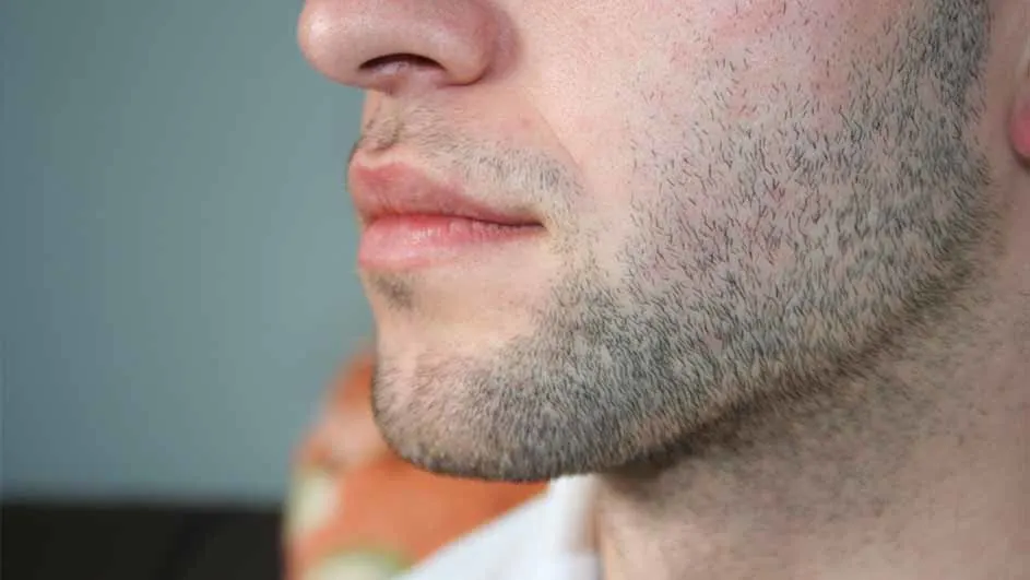 Profiel van een man met een korte baard, zichtbaar zijn neus, mond en kin tegen een grijze achtergrond.