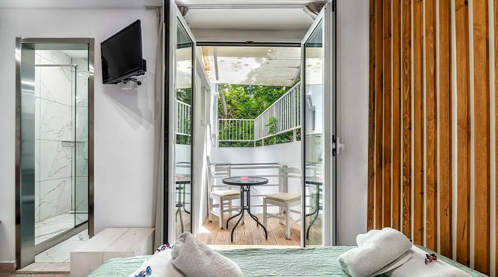 Slaapkamer met balkon, glazen douchecabine en houten wandpanelen. Buiten staan een tafel en stoelen. Een tv hangt aan de muur.
