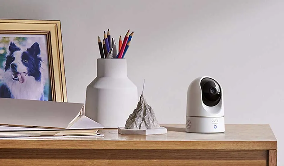 Op een houten bureau staat een witte Eufy beveiligingscamera naast een vaas met potloden, papieren en een foto van een hond.