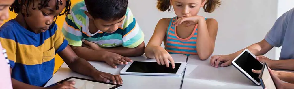 Meerdere kinderen zitten aan een tafel met tablets voor een gele en witte achtergrond.