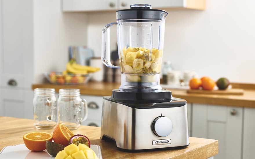 A glass blender jug filled with chopped banana and pineapple sits on a stainless steel base, beside jars and fresh fruit on a bright wooden worktop.