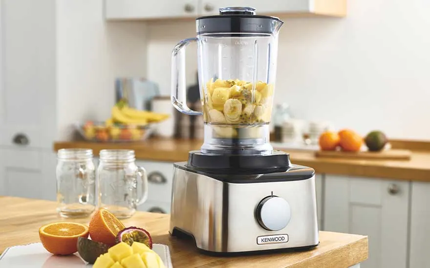 A glass blender jug filled with chopped banana and pineapple sits on a stainless steel base, beside jars and fresh fruit on a bright wooden worktop.