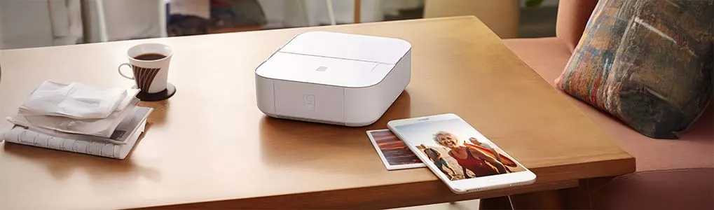 Een witte pocketprinter op een houten tafel naast een smartphone en geprinte foto, in een gezellige woonkamer met koffie en planten.