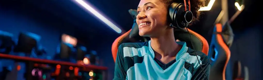 Een glimlachende persoon met een blauw-zwart shirt en een oranje koptelefoon zit in een gamestoel. Neonlichten op de achtergrond.