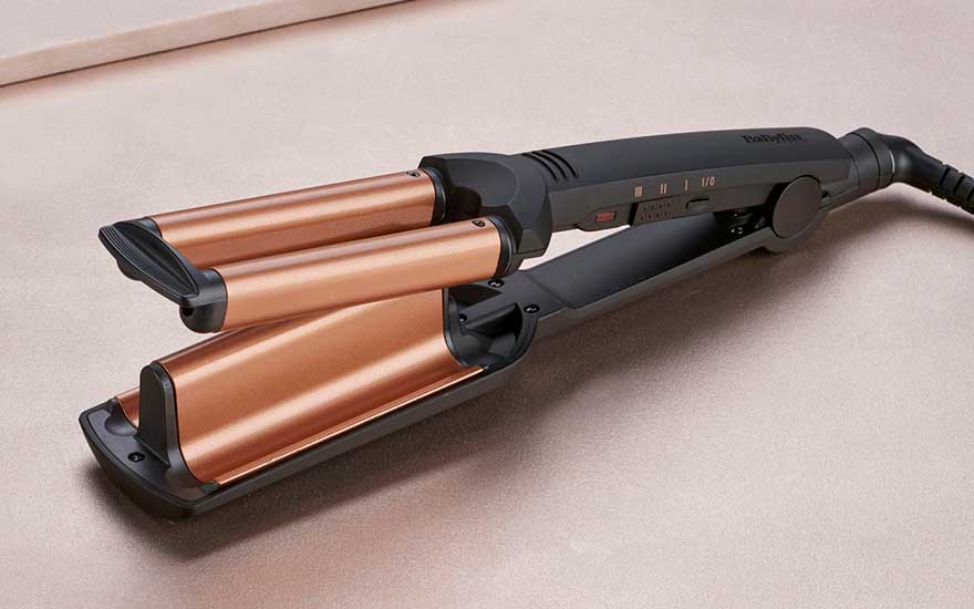 A hair styling tool with three copper-colored barrels and black handle, designed for creating waves, lying on beige surface.