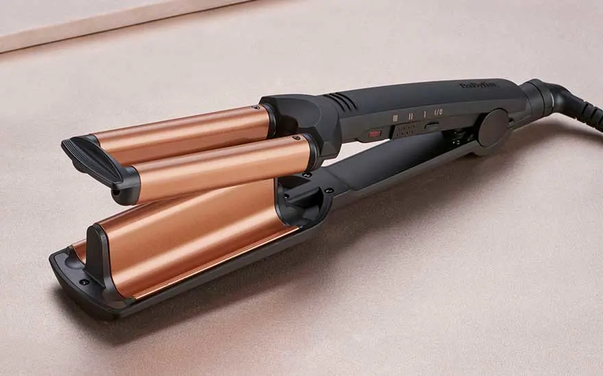 A hair styling tool with three copper-colored barrels and black handle, designed for creating waves, lying on beige surface.