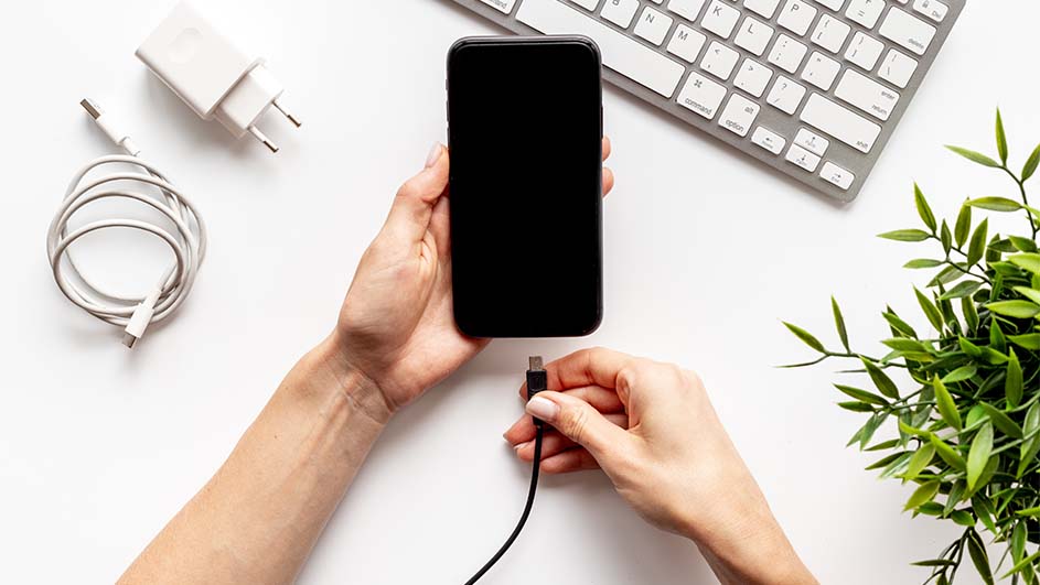 Twee handen verbinden een zwarte telefoon met een zwarte kabel. Een witte oplader, toetsenbord en plant zijn zichtbaar op een witte achtergrond.