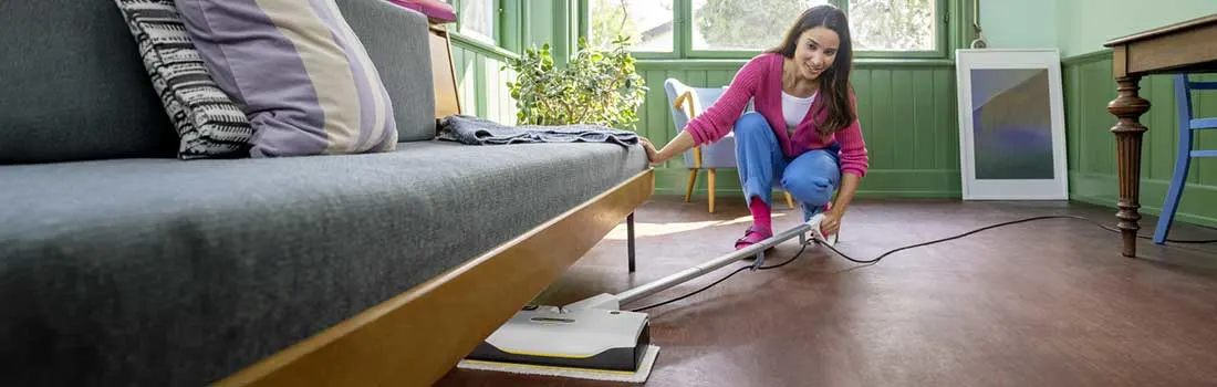 Vrouw schuift een platte stoomreiniger onder de bank in een lichte woonkamer en bereikt moeiteloos stof en vuil op moeilijk toegankelijke plekken.
