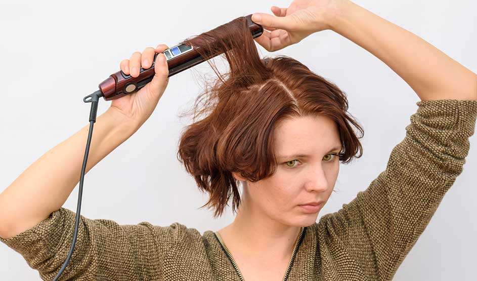 A woman with short auburn hair styles a section with a curling iron and looks focused against a plain background.