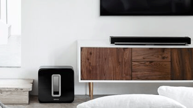 Zwarte subwoofer naast een houten kast met een witte rand en een zwarte soundbar erop, onder een zwarte tv.