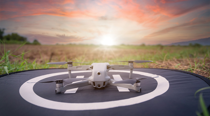 Een grijze drone staat op een zwart platform met een wit cirkelpatroon, tegen een zonsondergang en een veld.