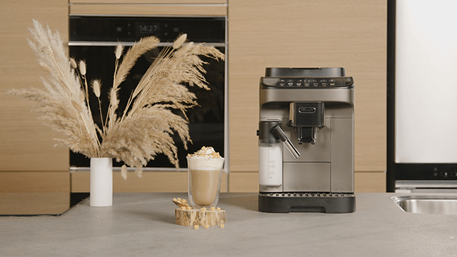 Een koffiezetapparaat naast een glas latte met slagroom, een vaas met pluimen en een oven in de achtergrond.