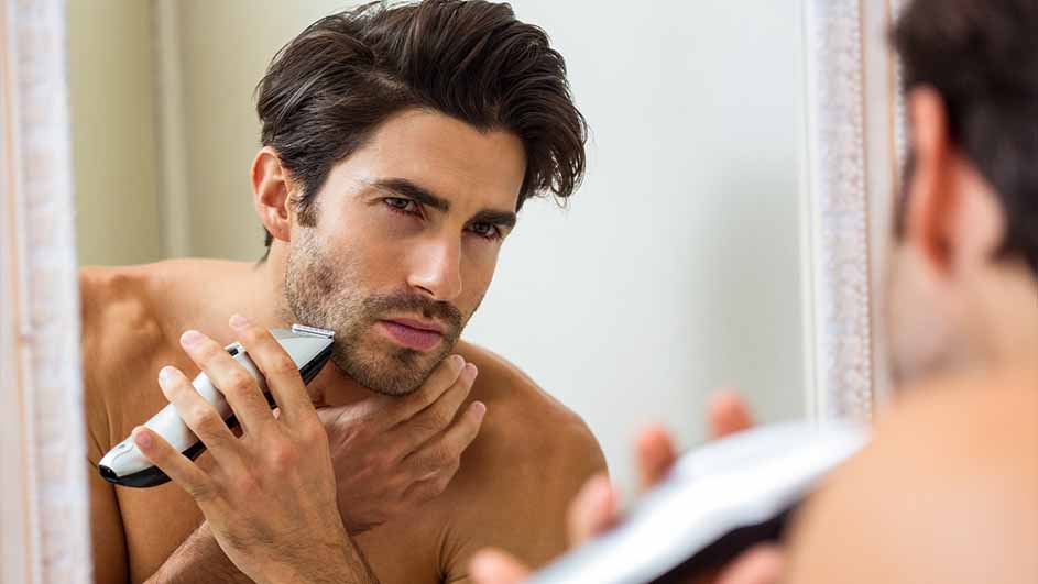 Een man staat voor een spiegel en gebruikt een witte trimmer om zijn baard te verzorgen in een lichte ruimte.