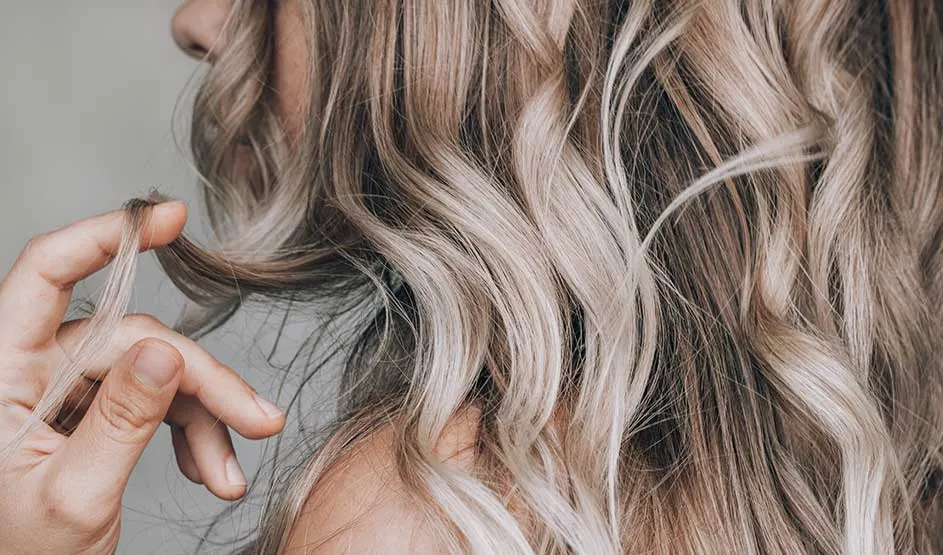 Long blonde locks fall in soft waves over one shoulder, while a hand playfully holds a strand of hair.
