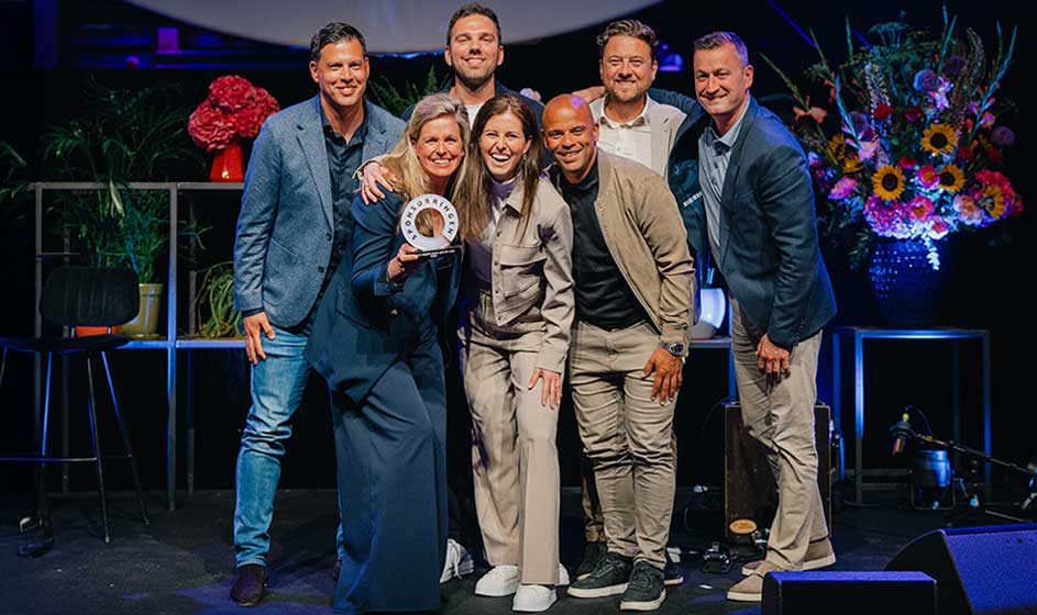 Groep van zes mensen poseert lachend op een podium; één persoon houdt een ronde prijs of trofee vast bij bloemen en licht.

