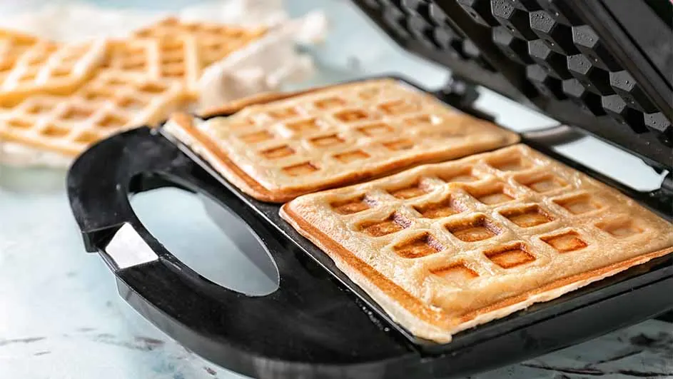 Twee goudbruine wafels in een zwart wafelijzer. Op de achtergrond liggen meer wafels op een lichtblauwe ondergrond.