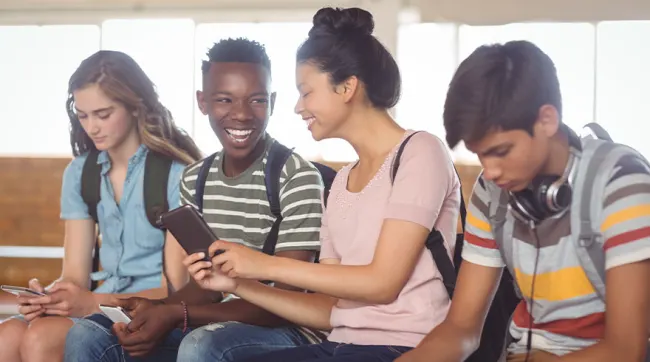 Vier tieners zitten, sommigen kijken naar telefoons. Ze dragen rugzakken en gestreepte shirts. Ramen op de achtergrond.