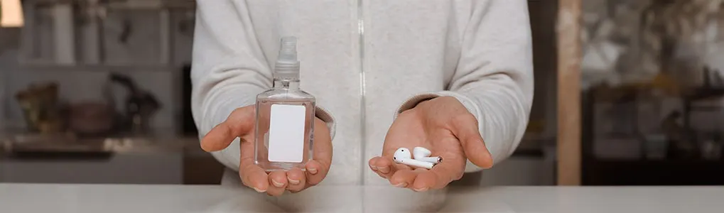 A person holds a hand sanitizer bottle and white earbuds. Background shows a shelf with objects.