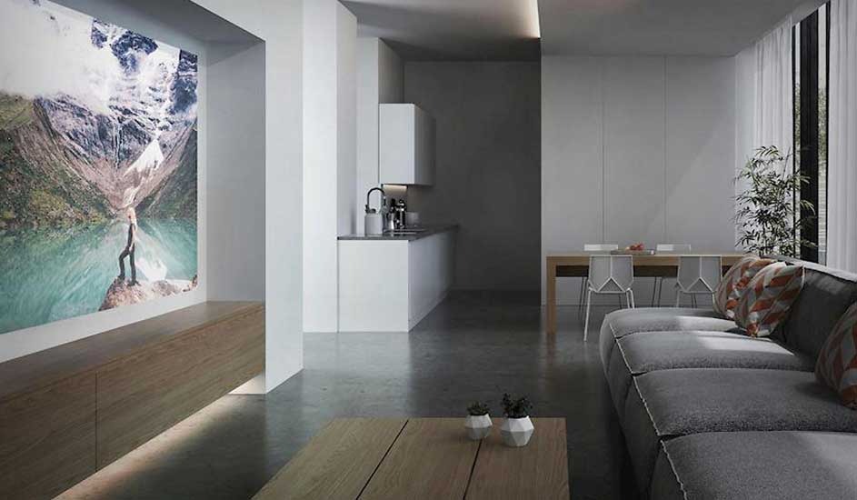 A modern living room with a grey sofa and wooden table features a large wall projection of a mountain and lake scene. No brand name is visible.