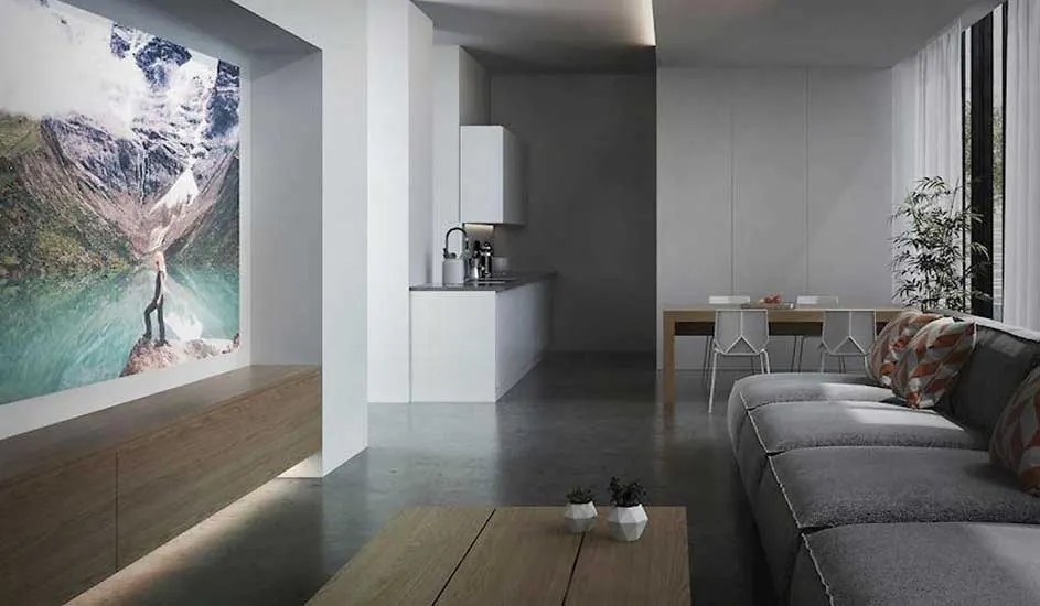 A modern living room with a grey sofa and wooden table features a large wall projection of a mountain and lake scene. No brand name is visible.