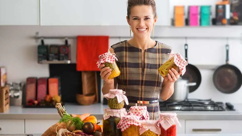 Vrouw in keuken met ingemaakte groenten in potten. Ze houdt twee potten vast, er staan er meer op het aanrecht.