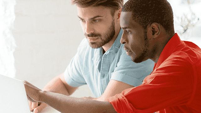 Twee mannen, een met een blauw overhemd en een met een rood overhemd, kijken naar een laptop.