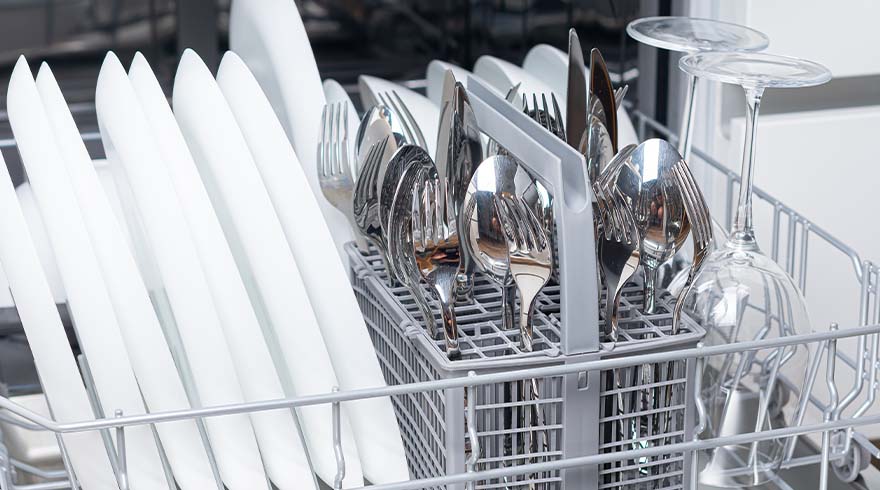 Een vaatwasserrek vol met zilveren lepels, vorken en messen, en twee glazen met een lange steel.