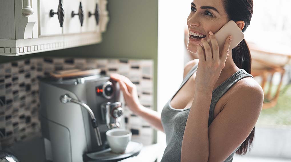 Een vrouw in een grijze tanktop lacht terwijl ze een telefoon vasthoudt en een koffiezetapparaat bedient in een keuken.