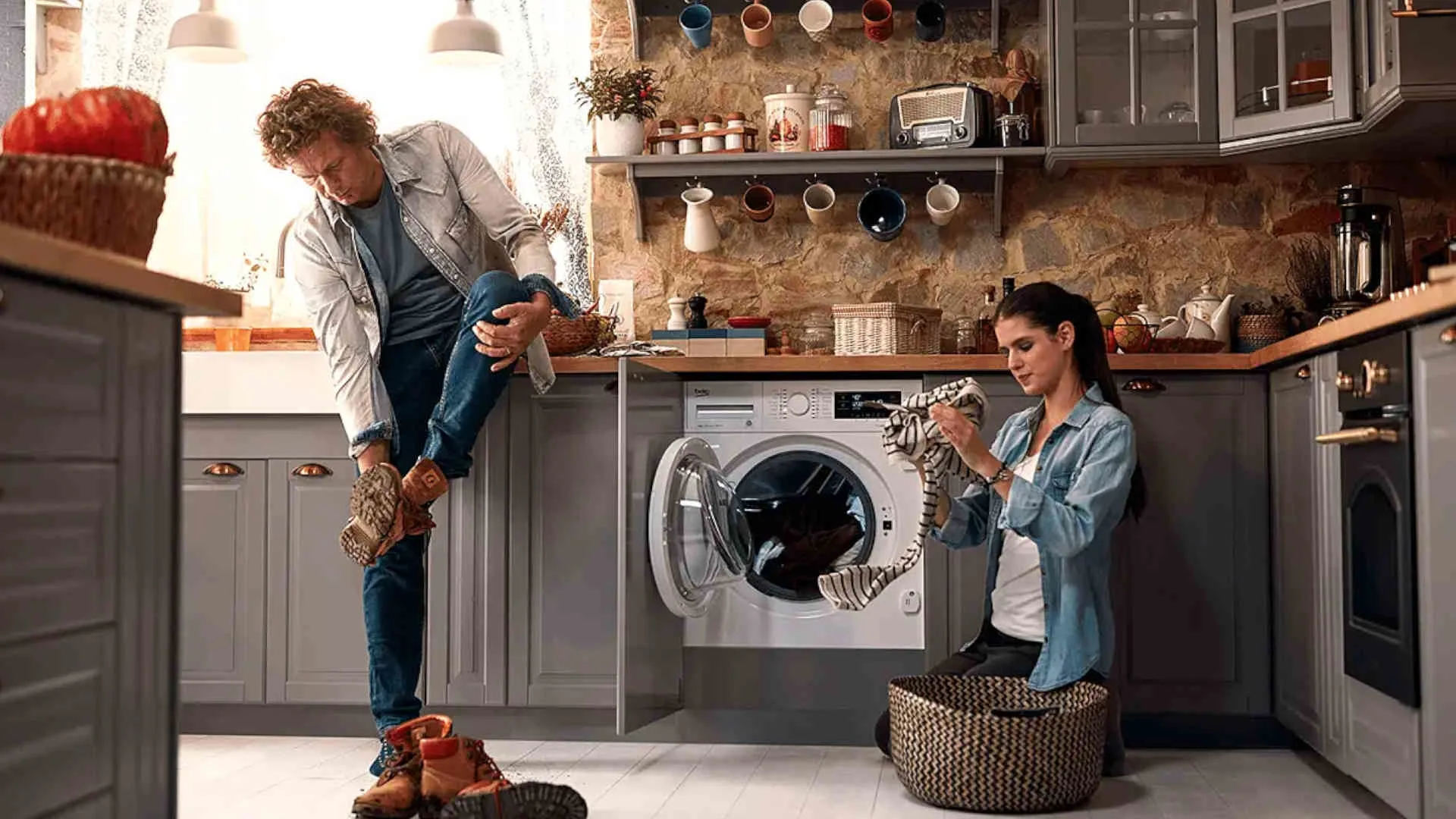 Een man trekt schoenen uit naast een wasmachine, terwijl een vrouw wasgoed sorteert in een keuken met grijze kasten.
