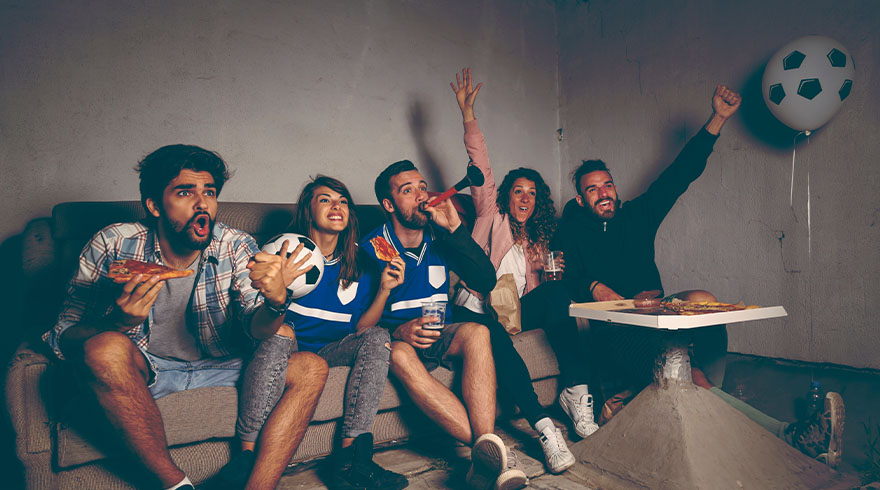 Vijf mensen zitten op een bank, kijkend naar een wedstrijd met pizza en een voetbalballon.