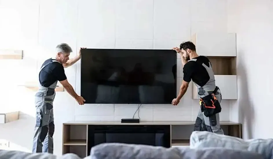 Two fitters are hanging a large flat-screen TV together on the wall above a modern TV unit in a bright living room.