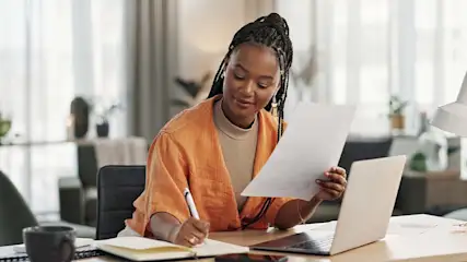 Een vrouw zit aan een bureau met een laptop en papieren, schrijvend in een notitieboek. Een kopje en decor staan op het bureau.