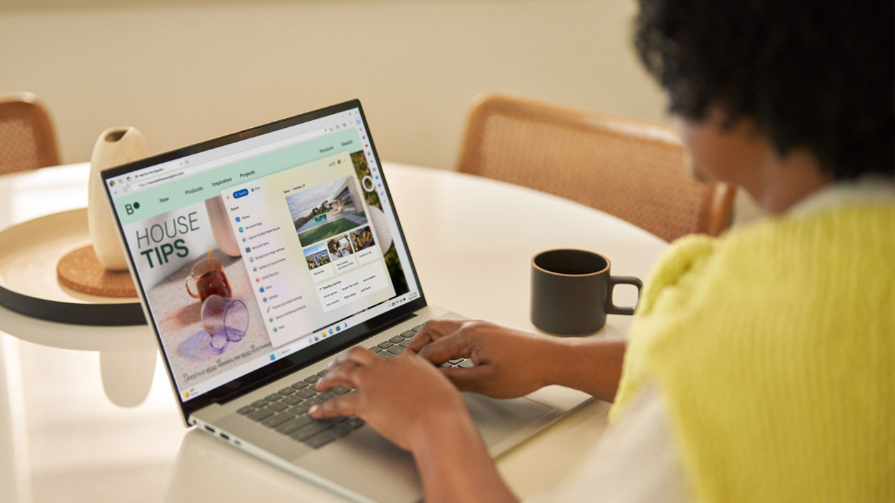 Iemand typt op een laptop op een witte tafel. Op het scherm staat "House Tips". Een mok en decoratie staan op tafel.