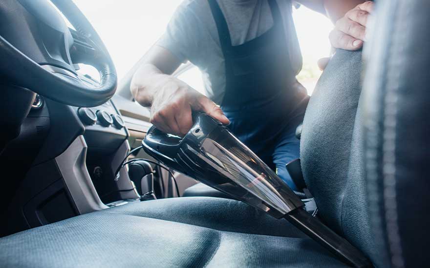 Iemand stofzuigt vakkundig de bestuurderstoel van een auto met een handstofzuiger, let goed op alle hoeken en blijft geconcentreerd.