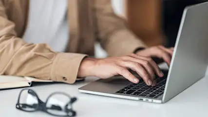 Iemand typt op een laptop; een notitieboek en een bril staan op een witte tafel.