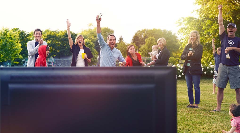 A group of people are gathered outside, watching a television screen and reacting with excitement, in a grassy area.