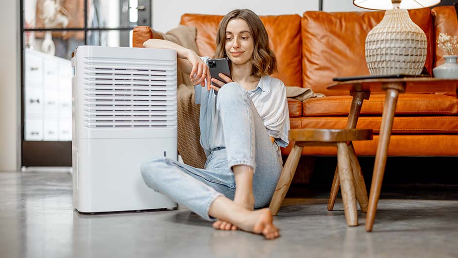 Vrouw zit op de grond met een telefoon, naast een witte luchtontvochtiger. Achter haar staat een oranje bank en een houten tafeltje.