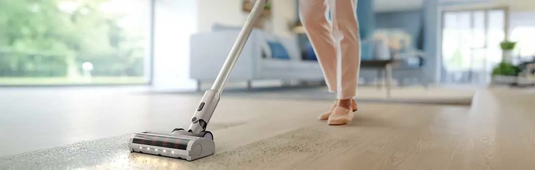 Iemand stofzuigt een houten vloer met een lichte steelstofzuiger, waarbij een strook kruimels netjes wordt opgezogen in de zonnige woonkamer.