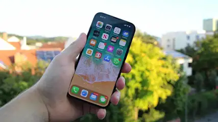 Een hand houdt een smartphone met een scherm vol app-pictogrammen en een strandachtergrond vast. Op de achtergrond zijn bomen en gebouwen te zien.