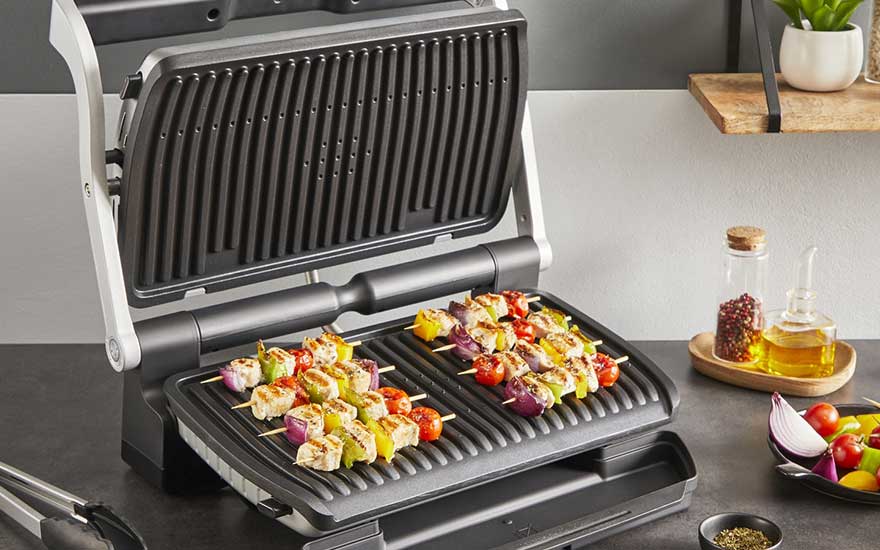 An electric contact grill sizzles with colourful chicken and vegetable skewers, while herbs, oil and fresh veg stand ready beside it.