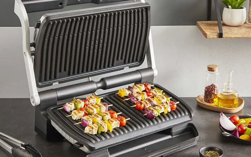 An electric contact grill sizzles with colourful chicken and vegetable skewers, while herbs, oil and fresh veg stand ready beside it.