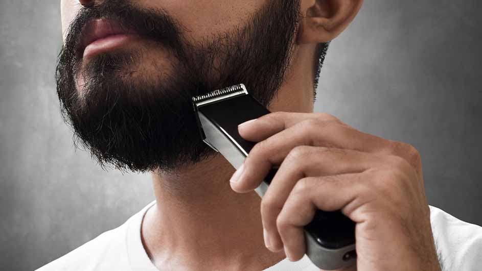 Een man trimt zijn baard met een zwarte tondeuse tegen een grijze achtergrond.