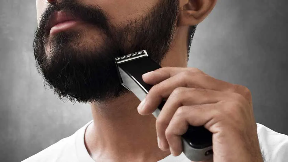 Een man trimt zijn baard met een zwarte tondeuse tegen een grijze achtergrond.