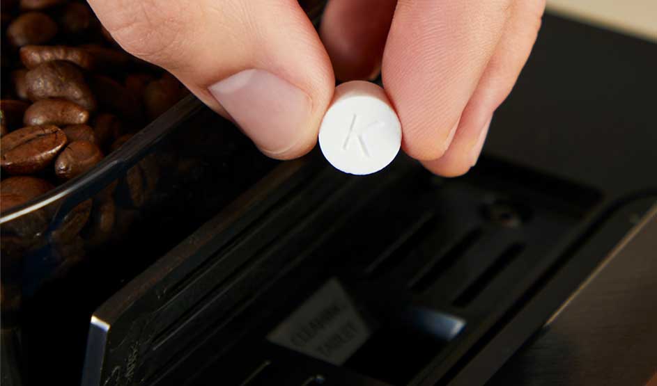 Een hand plaatst een reinigingstablet met de letter K in een koffiemachine naast koffiebonen. Het merk is niet zichtbaar op deze afbeelding.