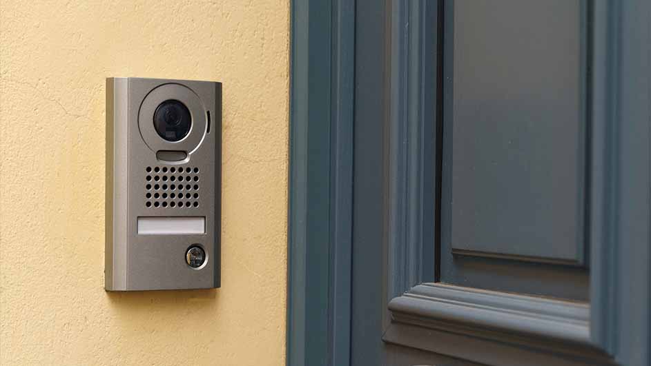 Metalen intercom met camera aan een gele muur, naast een blauwe deur.