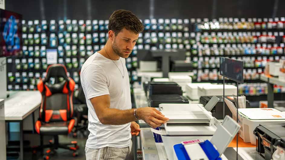 Een man in een winkel bekijkt een printer op een toonbank met andere printers; op de achtergrond staan rekken vol producten.