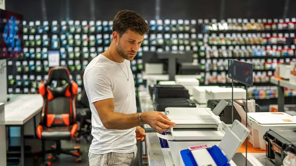Een man in een winkel bekijkt een printer op een toonbank met andere printers; op de achtergrond staan rekken vol producten.
